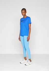Sininen Nike-paita, jossa on musta swoosh-logo, yhdistettynä vaaleansinisiin legginsseihin ja valkoisiin urheilukenkiin, harmaata taustaa vasten.