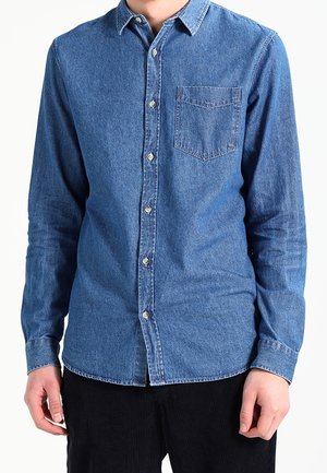 Mężczyzna w niebieskiej, długorękawowej koszuli jeansowej zapinanej na guziki, z kieszenią na piersi i czarnych spodniach, stoi na białym tle.
