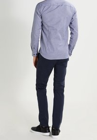 Man wearing a blue checkered shirt, dark navy pants, and black sneakers standing, shown from the back against a plain background.