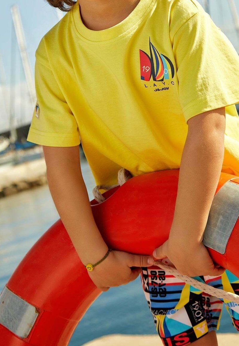 Enfant portant une chemise de voile jaune et un short coloré tient une bouée rouge près de l'eau avec des mâts de bateaux en arrière-plan.