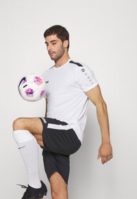 Maglietta atletica bianca con dettagli neri, pantaloni corti neri e calze bianche. Un pallone da calcio è bilanciato su un ginocchio. Design semplice, materiale traspirante.