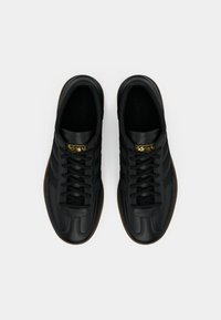 Μαύρα δερμάτινα αθλητικά παπούτσια με υφασμάτινες πλευρές, διαθέτοντας τρεις μαύρες ρίγες, χρυσό λογότυπο και σόλα από καουτσούκ τύπου γκαμ. Κλασικός σχεδιασμός με κορδόνια.