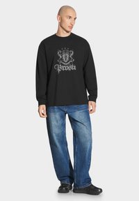 Czarny sweter z długim rękawem z szarym wzorem graficznym i tekstem z przodu, połączony z luźnymi niebieskimi dżinsami i czarnymi butami.