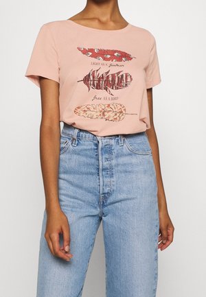 Persona que lleva una camiseta rosa con tres plumas estilizadas y el texto "Light as a feather, free as a bird", metida en unos vaqueros azules de talle alto.