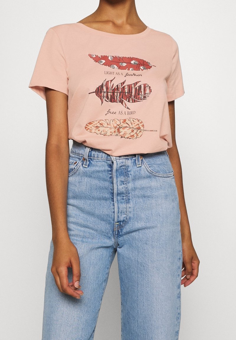 Personne portant un t-shirt rose avec trois plumes stylisées et le texte "Light as a feather, free as a bird", rentré dans un jean bleu taille haute.