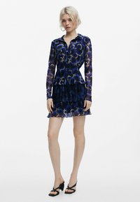 Langärmliges blaues Kleid mit Blumenmuster, Falten-Design und geschichteter Rock. Kombiniert mit schwarzen Absatzsandalen.