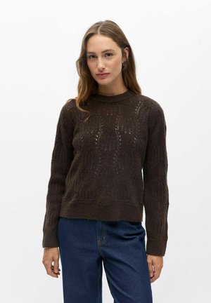 Femme aux longs cheveux bruns portant un pull en tricot brun foncé et un jean bleu, debout devant un fond blanc uni.