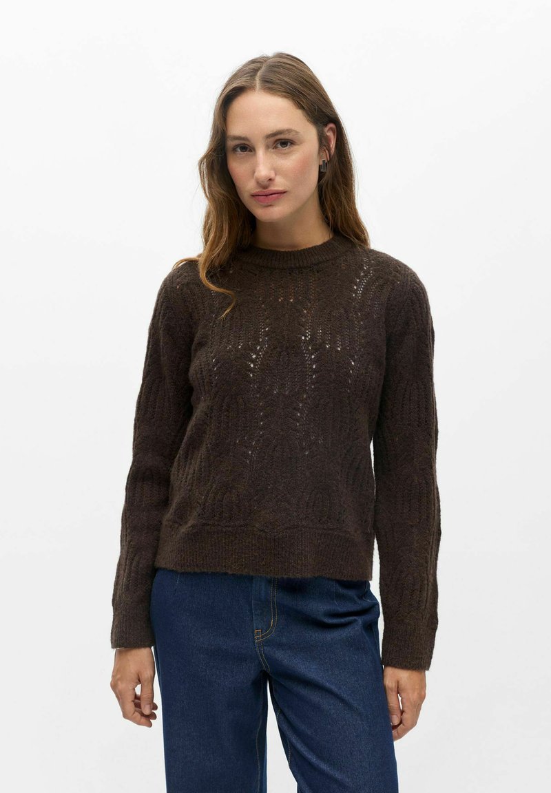 Femme aux longs cheveux bruns portant un pull en tricot brun foncé et un jean bleu, debout devant un fond blanc uni.