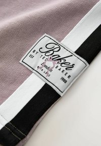 Lyse lilla stof med hvide og sorte striber; har et mærkat med teksten "Baker by Ted Baker", lyserødt sløjfe-design og etableringsdatoen 1988.