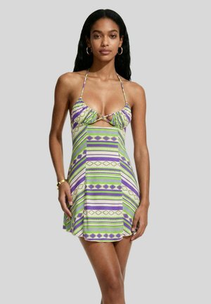 Donna che indossa un mini abito senza maniche con scollo halter e motivi geometrici verdi, viola e crema, in piedi davanti a uno sfondo semplice.