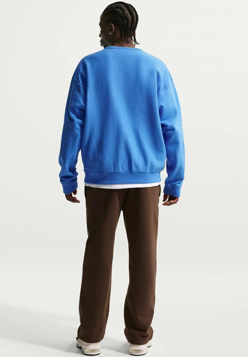 Mann mit geflochtenem Haar, der einen blauen Pullover, braune Hosen und weiße Turnschuhe trägt, steht mit dem Rücken zur Kamera vor einem einfarbigen Hintergrund.