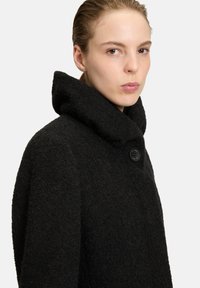 Zwarte wollen jas met een hoge kraag, gestructureerde stof en een enkele knoopsluiting, die een gestructureerd, minimalistisch ontwerp laat zien.