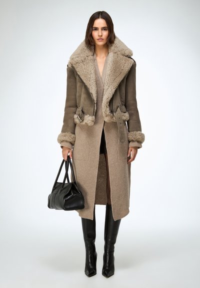 Femeie purtând o rochie tricotată bej, o jachetă maro de blană, cizme negre peste genunchi, ținând o geantă de mână din piele neagră, stând în picioare pe fundal alb.