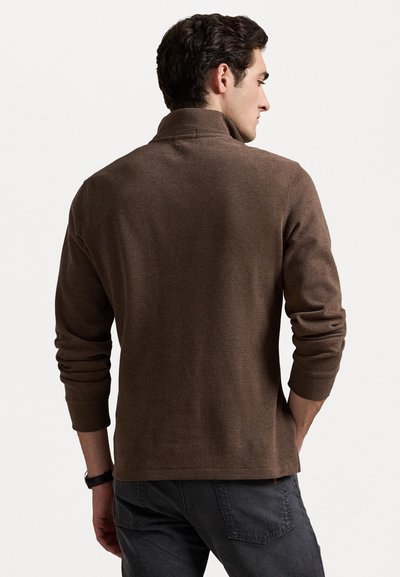 Pull en maille côtelée marron avec un col haut et des manches longues, présentant une coupe décontractée et un tissu texturé. Détails de couture discrets.