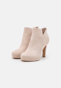 Un par de botines de ante beige con tacones altos y una sutil plataforma sobre un fondo blanco.