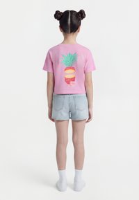 Παιδί με χτένισμα διπλό κότσο, φοράει ροζ κομμένο μπλουζάκι με γραφικό κομμένης ανανά και ανοιχτόχρωμα σορτς τζιν, κοιτάζει προς τα πίσω.