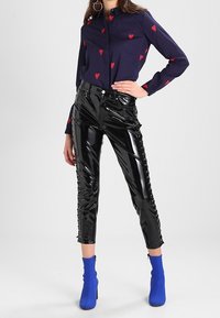 Marineblaues Hemd mit verstreuten roten Herzmotiven; glänzende schwarze Lacklederhosen mit seitlicher Schnürung; blaue Knöchelstiefel.