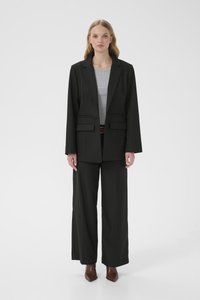 Fekete, testre szabott blézer két első zsebbel, széles szárú fekete nadrággal párosítva. A figurán szürke felső és barna hegyes orrú magassarkú cipő látható.