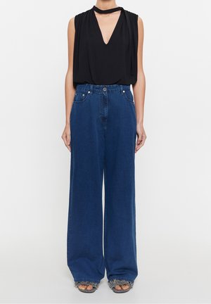 Person iført sort ærmeløs V-hals top med nakkesnøre og højtaljede, vidtbenede blå jeans med frynsekanter og sandaler.