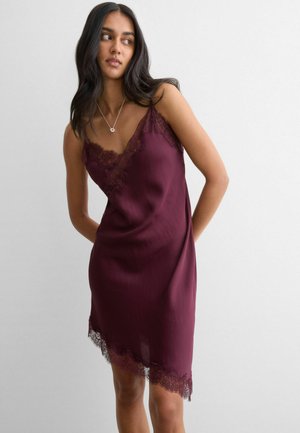 Frau mit langem dunklem Haar, die ein ärmelloses burgunderrotes Nachthemd mit Spitzenbesatz trägt und vor einem schlichten hellen Hintergrund steht.