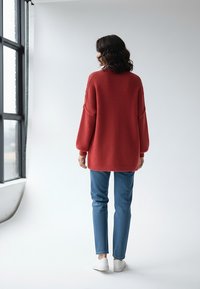 Za duży czerwony dzianinowy sweter z opuszczonymi ramionami, zestawiony z niebieskimi dżinsami i białymi trampkami, na jednolitym jasnym tle.