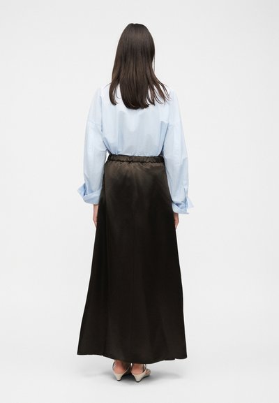 Γυναίκα με μακριά σκούρα μαλλιά που φοράει ανοιχτό μπλε πουκάμισο, μακριά μαύρη φούστα και μπεζ τακούνια, στέκεται με την πλάτη σε απλό φόντο.