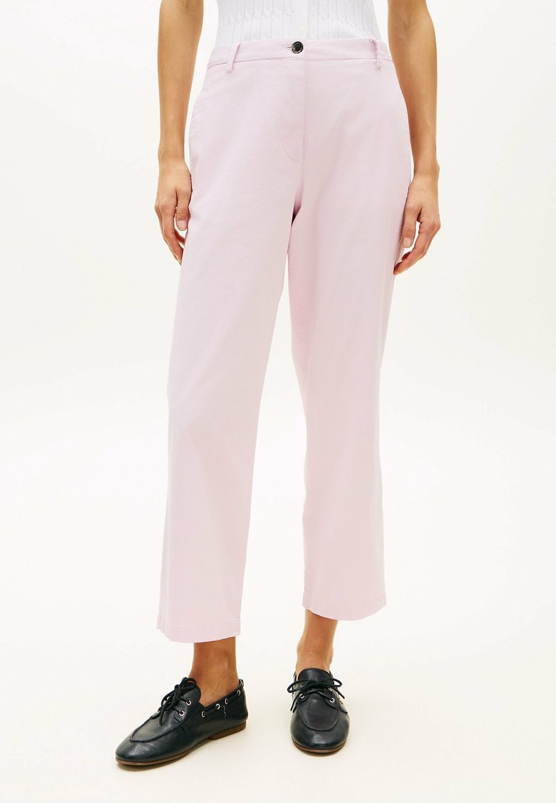 Personne portant un pantalon rose clair longueur cheville, des chaussures noires à lacets et un haut blanc côtelé, debout devant un fond uni.