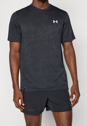 Homme portant un t-shirt noir à manches courtes Under Armour et un short noir Project Rock, debout devant un fond uni.