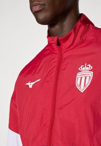 Červená ľahká bunda s predným zipsom, bielymi akcentmi a logami AS Monaco a Mizuno na hrudi. Hladká textúra.