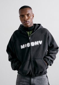 Schwarze Zip-up-Hoodie mit einem auffälligen "MISBHV"-Logo in Weiß, ausgestattet mit einer Kapuze und elastischen Bündchen, getragen über einem gestreiften Oberteil und grauen Jeans.