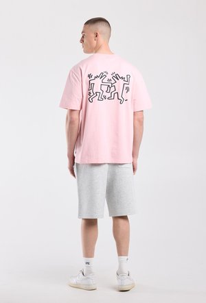 Man die van achteren is gefotografeerd, draagt een roze T-shirt met illustratie van zwarte dansfiguren, grijze korte broek, witte sokken en witte sneakers.