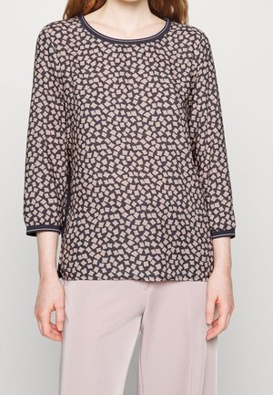 Langærmet top med et geometrisk mønster af beige og mørkebrune firkanter, rund halsudskæring og sort kant, lavet af letvægtsstof.