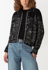 Svart cardigan med broscherade vita urbana mönster, knappstängning och klassisk krage. Bärs över en svart skjorta med blå denimjeans.