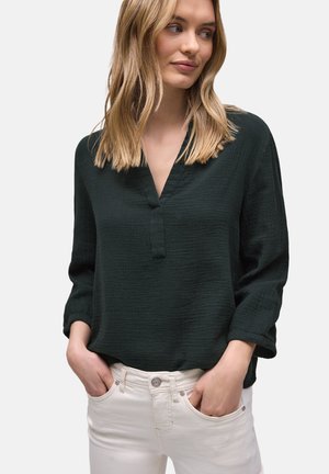 Femme blonde portant un chemisier à manches longues texturé vert foncé avec un décolleté en V et un pantalon blanc, mains dans les poches, regardant sur le côté.
