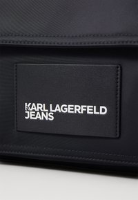 Juoda audinio maišas su nuošlifuota tekstūra ir iškilnia logotipo etikete, kurioje rašoma "KARL LAGERFELD JEANS" kontrastingai balta spalva.