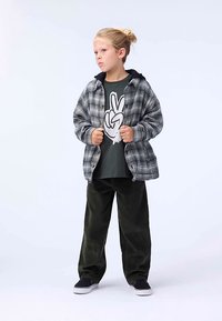 Flanelasta srajca v sivo-črni karo vzorcu, oblečena čez temno zeleno majico z grafiko. Ohlapne zelene hlače iz korduroja in črni supergi.