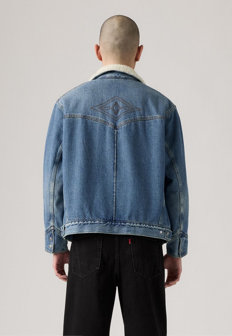 Lekka niebieska kurtka dżinsowa z kołnierzem z owczej wełny, haftowanym wzorem diamentu na plecach oraz zapięciem na guziki. Połączona z czarnymi dżinsami.