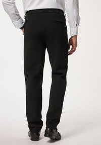Zwarte broek van een gladde stof, met een getailleerde pasvorm, zijzakken en een enkele achterzak. Draagt zwarte schoenen.