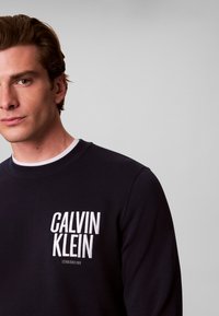 Jeune homme aux cheveux châtain clair portant un sweat Calvin Klein noir avec un logo blanc sur la poitrine sur un fond gris uni.