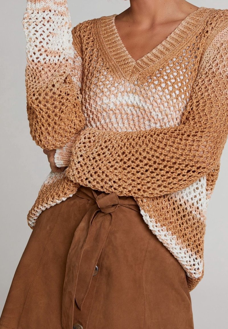 Pull en tricot marron avec col en V et design à maille ouverte, avec des accents de couleur plus claire. Associé à une jupe en daim marron nouée à la taille.