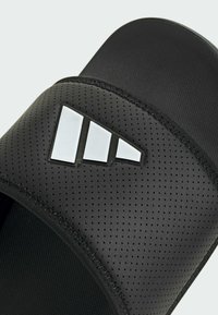 Black slide sandal featuring a textured matte strap with perforations and a white three-stripe logo on the front. Flat sole design.