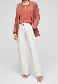 Blusa corallo con rifiniture in pizzo, abbinata a un blazer rosa chiaro e pantaloni bianchi a gamba larga. Sandali con tacco neutri completano il look.