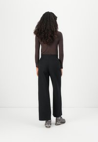 Black wide-leg pants with a high waistband, smooth texture, and two back pockets, paired with grey sneakers. Model has curly hair.