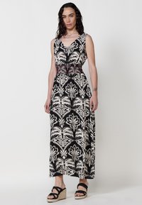 Robe maxi à motifs floraux noir et crème avec un décolleté en V, une taille cintrée et une ceinture tressée détaillée. Associée à des sandales compensées noires.