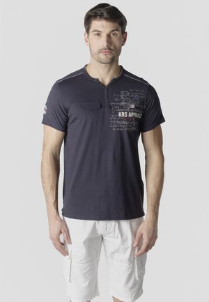 Hombre adulto con cabello corto y oscuro, que lleva una camiseta gráfica azul marino y pantalones cortos cargo blancos, de pie frente a un fondo liso.