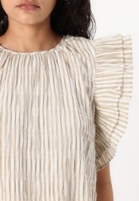 Haut clair avec des plis verticaux et des rayures beiges et blanches. Les manches volantes ajoutent de la texture ; le col rond améliore le design.