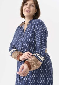 Niebieska sukienka w wzory z guzikiem z przodu, kontrastowym wzorzystym kołnierzykiem i teksturowanymi brązowymi mankietami. Gładki materiał, dopasowany krój.