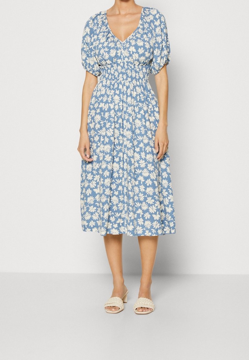Blaues Blumenkleid mit weißen Gänseblümchenmustern, kurzen Puffärmeln, einem V-Ausschnitt und einem elastischen Bund, kombiniert mit hellen Sandalen.