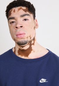 Námořnická modrá mikina s kulatým výstřihem, měkká tkanina, se známým bílým logem Nike na levé části hrudi. Detailní záběr osoby s vitiligem.
