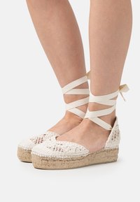 White crochet espadrilles with jute platform, featuring crisscross ankle ties. Open-toe design and textured fabric on the upper.